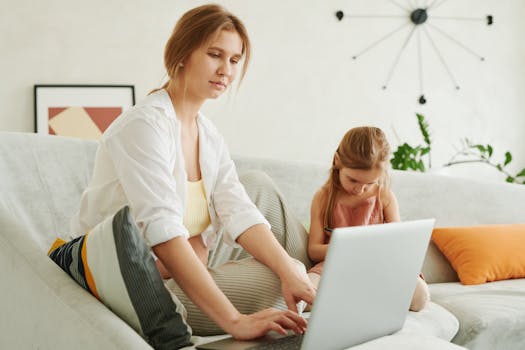 A mother and daughter use a laptop together on a cozy sofa indoors, multitasking family life.
